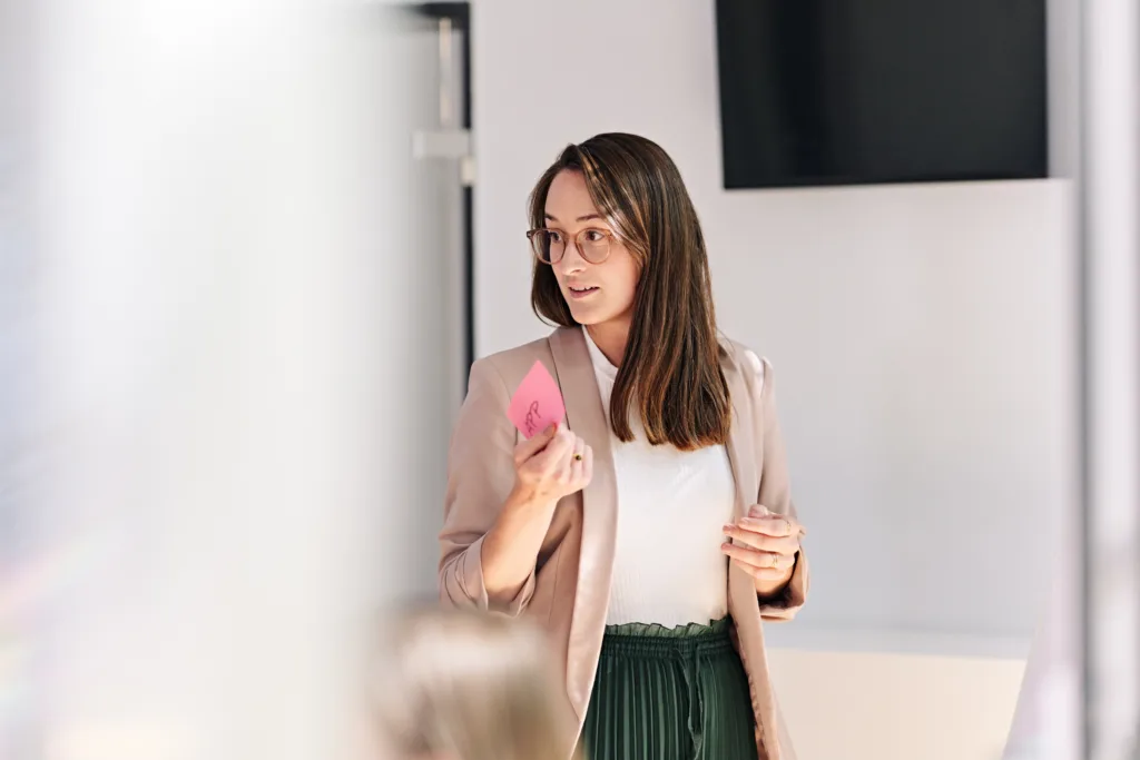 Eine Frau mit Brille und langen braunen Haaren, Vertreterin der KI Agentur, steht in einem Büro und hält einen rosa Haftzettel in der Hand. Sie trägt einen beigen Blazer und eine grüne Hose und blickt zur Seite. Im Hintergrund ist ein Flachbildfernseher zu sehen, der Vordergrund ist unscharf.